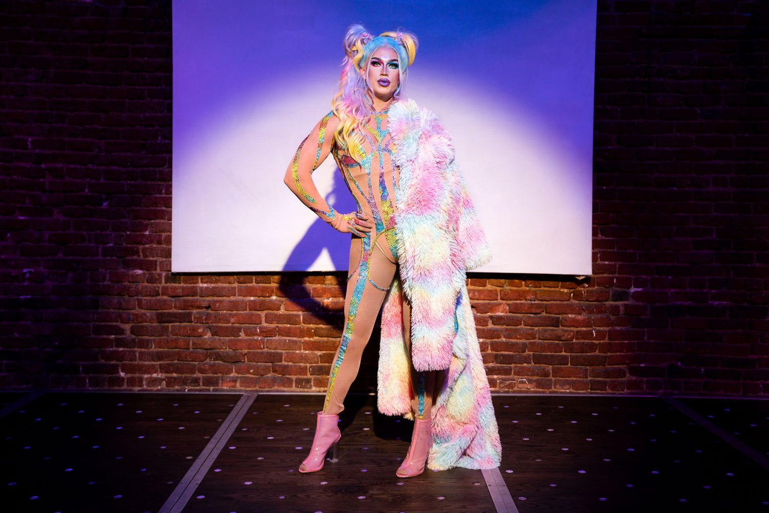A drag performer in a colorful pastel sequined outfit and feathered coat poses on a stage in front of a brick wall.