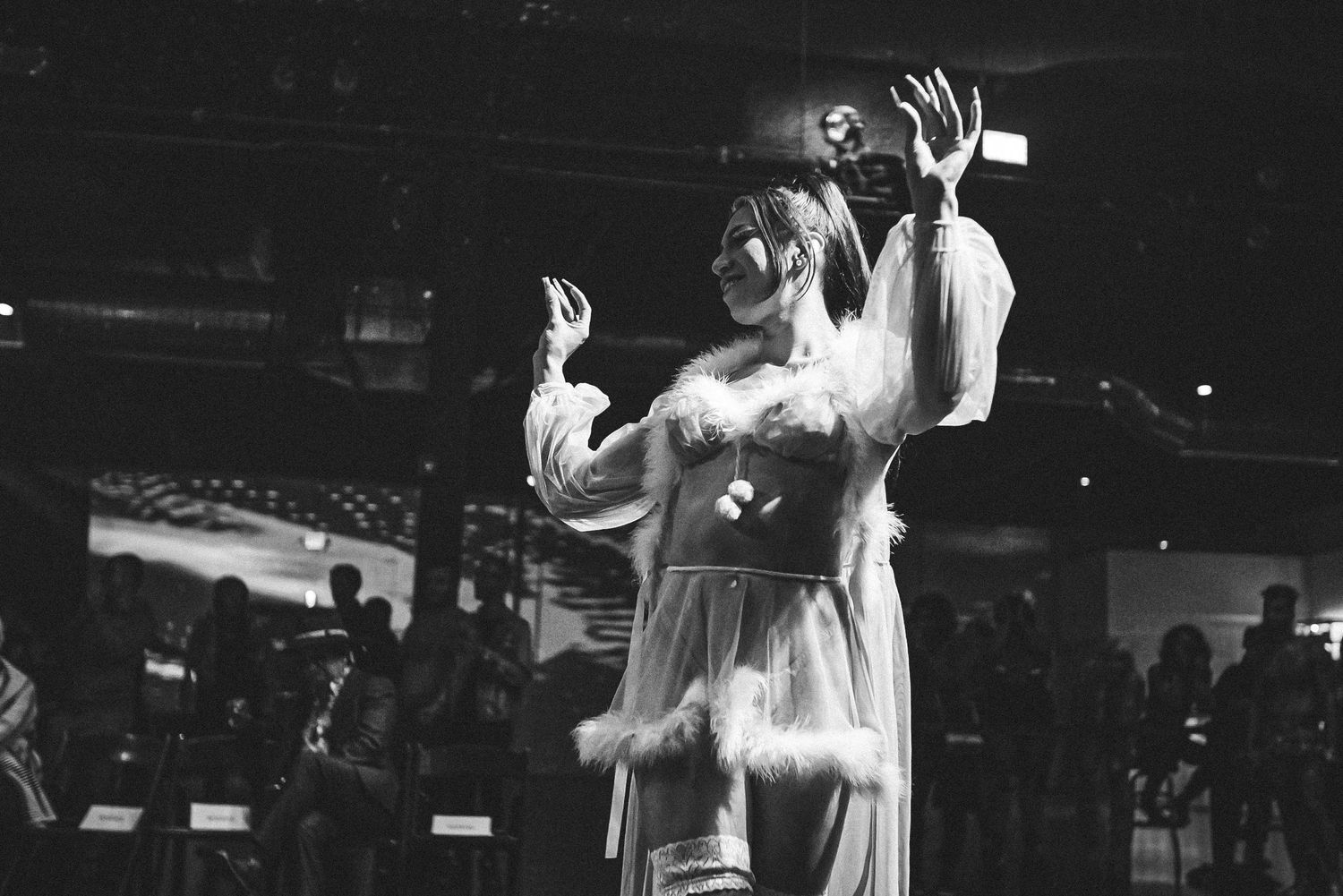 Black and white photo of dancer in white feathered costume performing with raised hands.