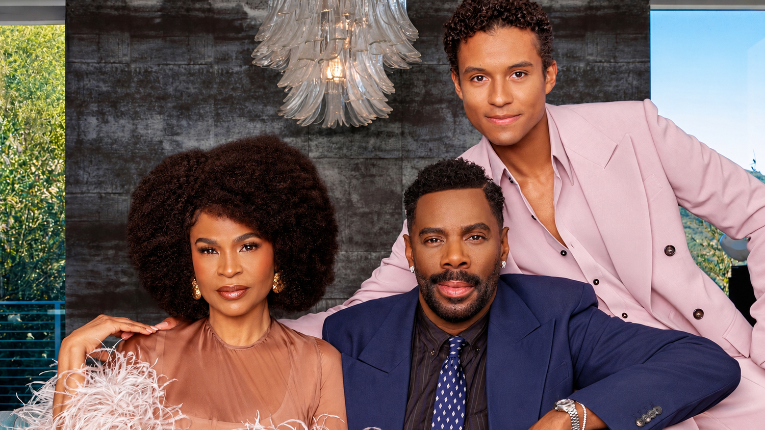 Three stylishly dressed people pose closely together in a modern room, a woman with a voluminous afro and two men in pink and blue suits.