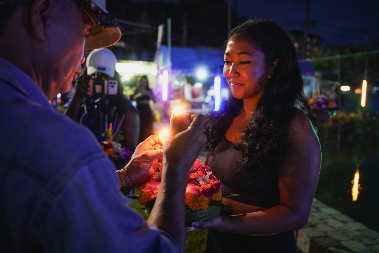A woman holding flowers receives a candle light during a nighttime cultural ceremony.