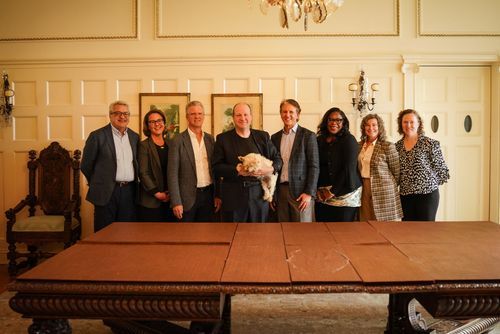 Group of seven professionals pose together around antique wooden table in elegant formal room.