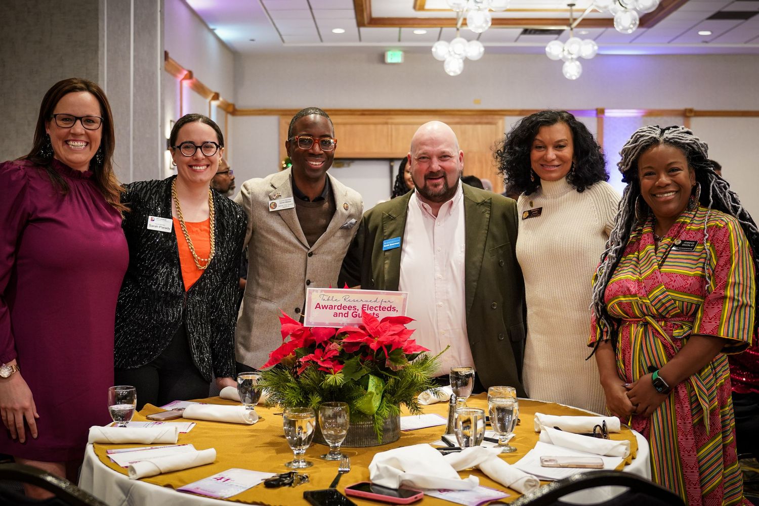 Six diverse professionals smile together at formal dinner event with holiday poinsettia centerpiece.