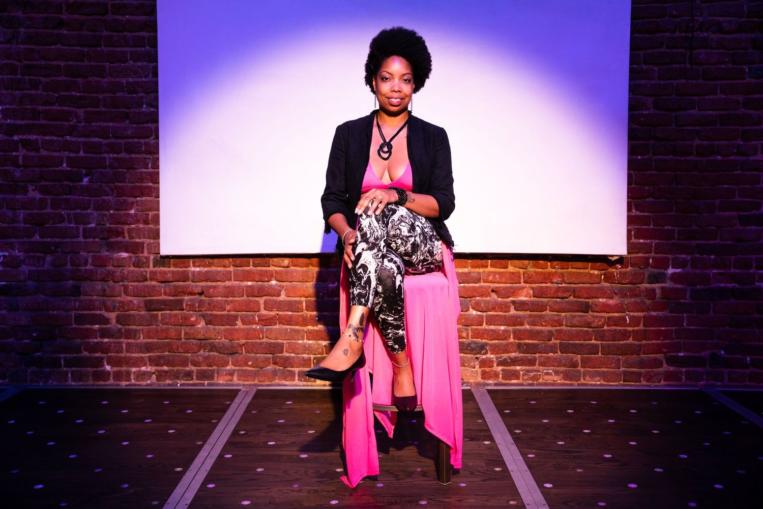 Stylish woman in a black blazer and pink pants posing confidently on a stage with a brick wall backdrop.