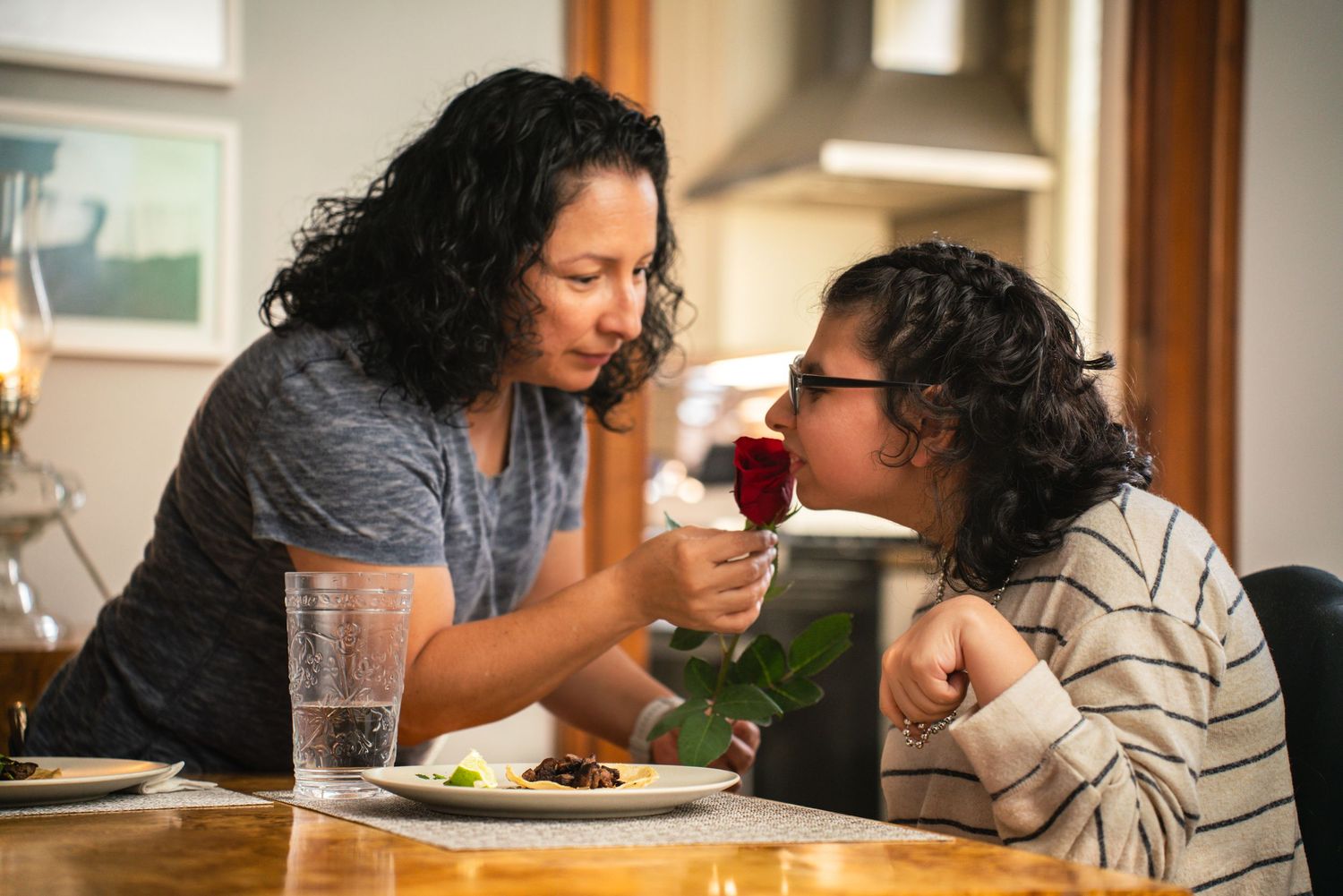 Mother feeding child wearing glasses a bite of food at wooden dining table at home.