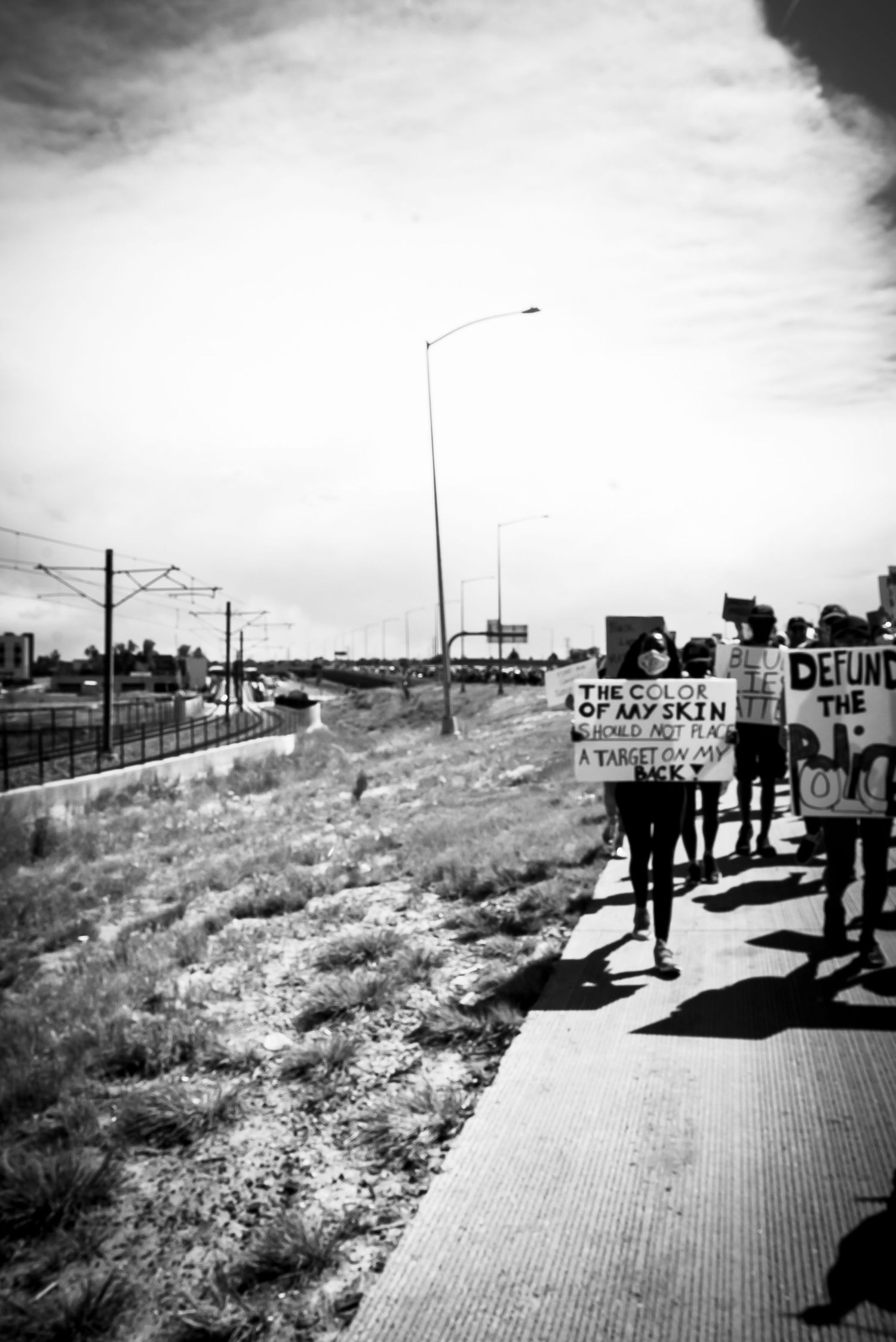 Black and white photo of protesters marching along a highway holding signs reading Defund The Police.