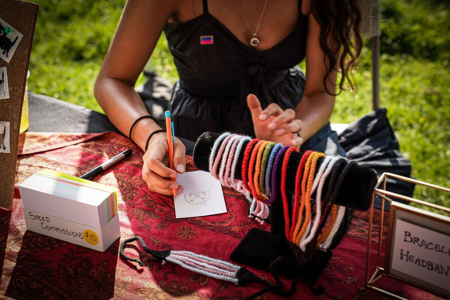 Person arranging colorful bracelets on a red cloth at an outdoor Pride vendor table.