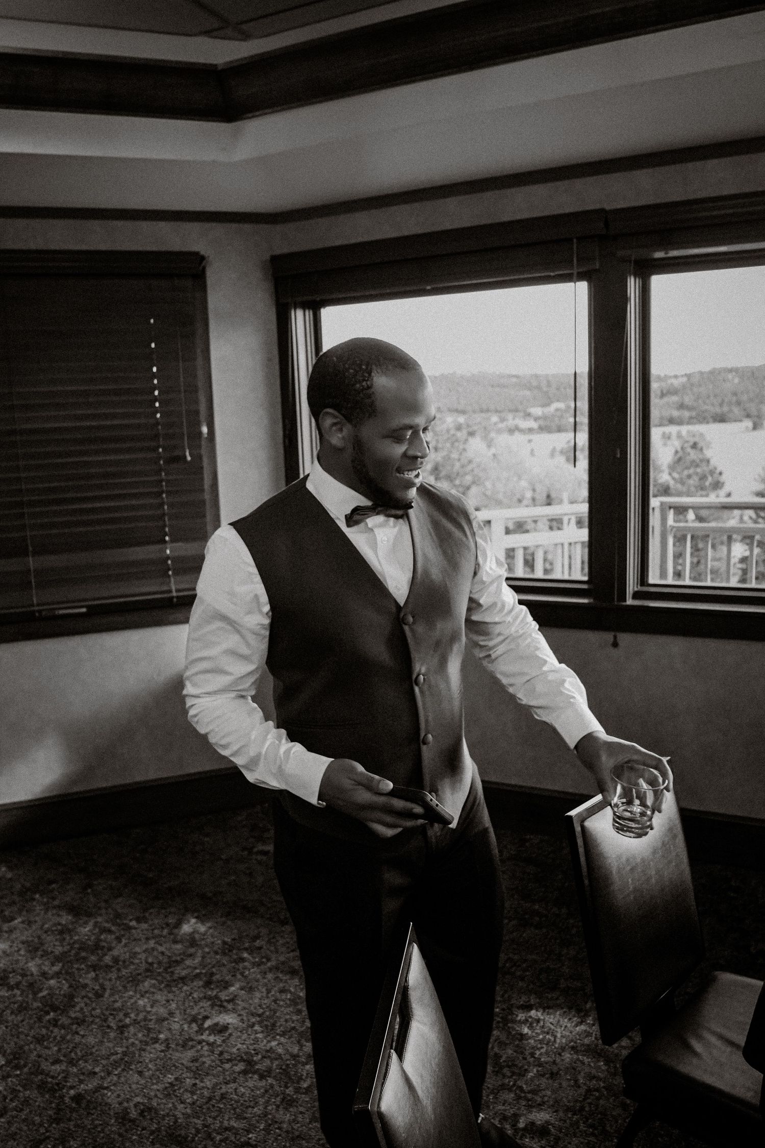 Black and white photo of a well-dressed groom in vest and bow tie holding a bag while standing near a window.