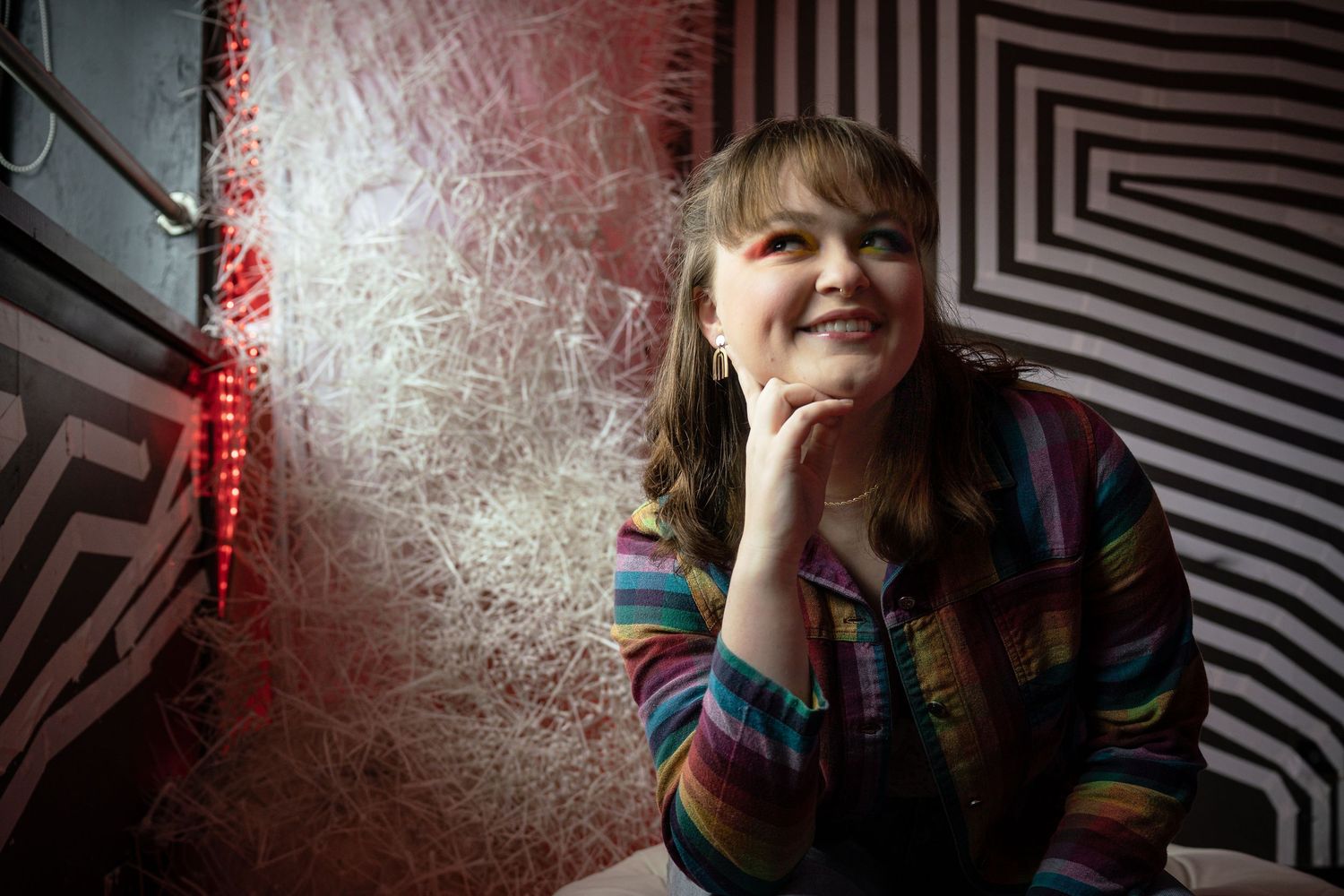 A smiling woman in a colorful striped shirt sits relaxed near a decorative textured wall with geometric black and white patterns.