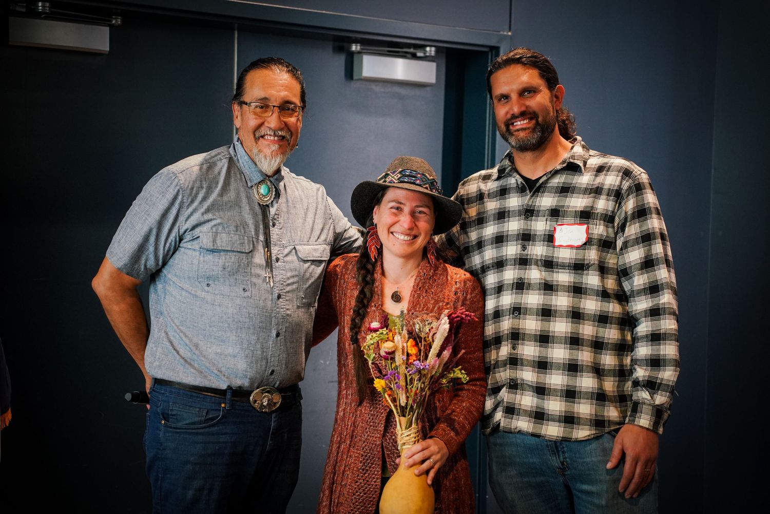 Three smiling people pose together indoors, woman in center holding colorful bouquet of autumn flowers.