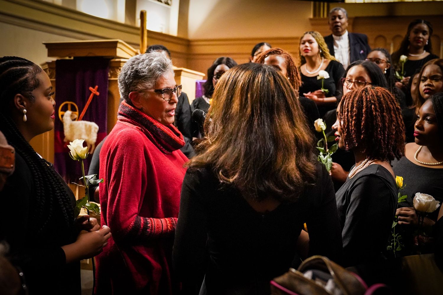 Diverse group of people in formal attire socializing and holding white roses inside a church.