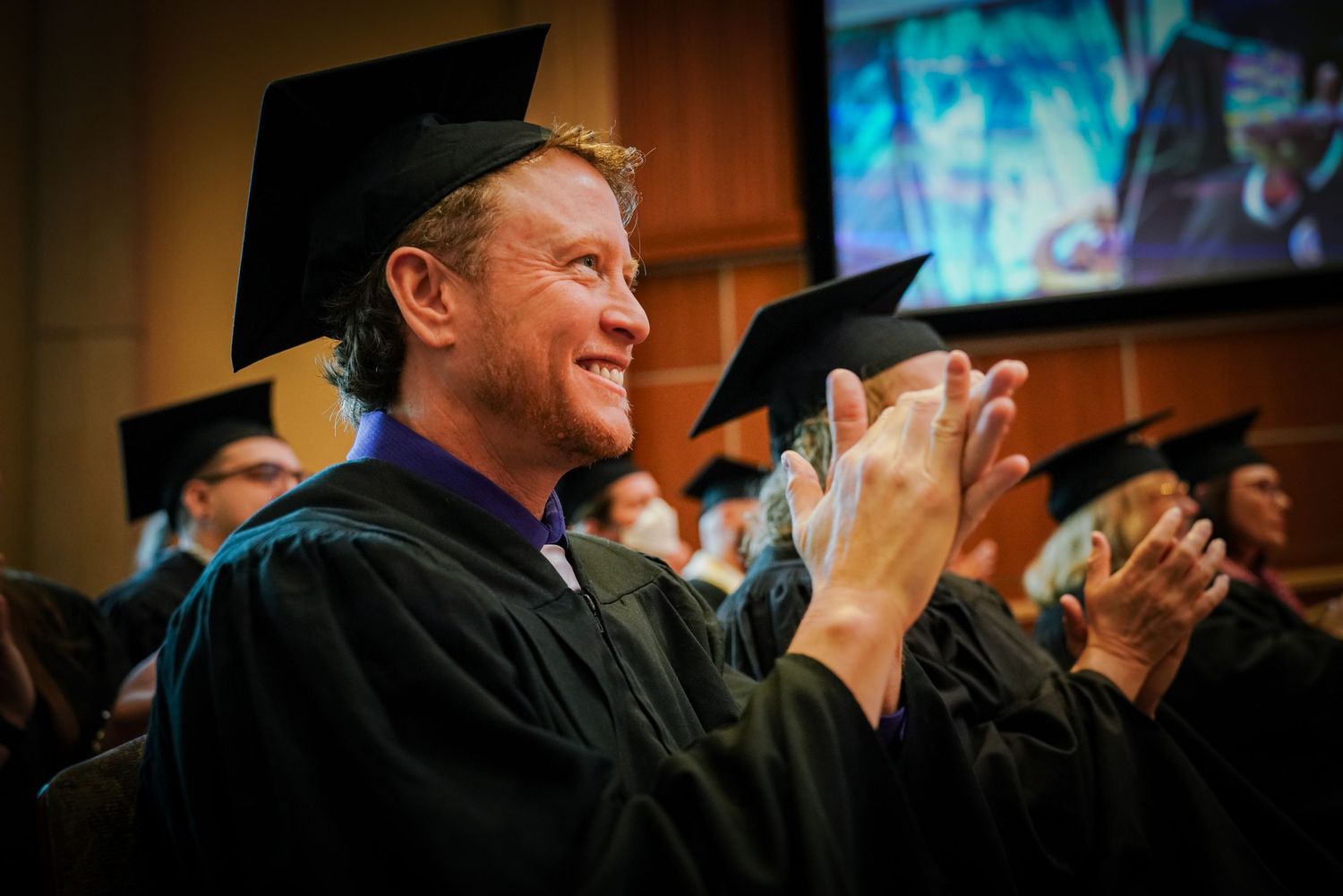 Smiling graduate in cap and gown applauds during ILIFF School of Theology commencement ceremony.