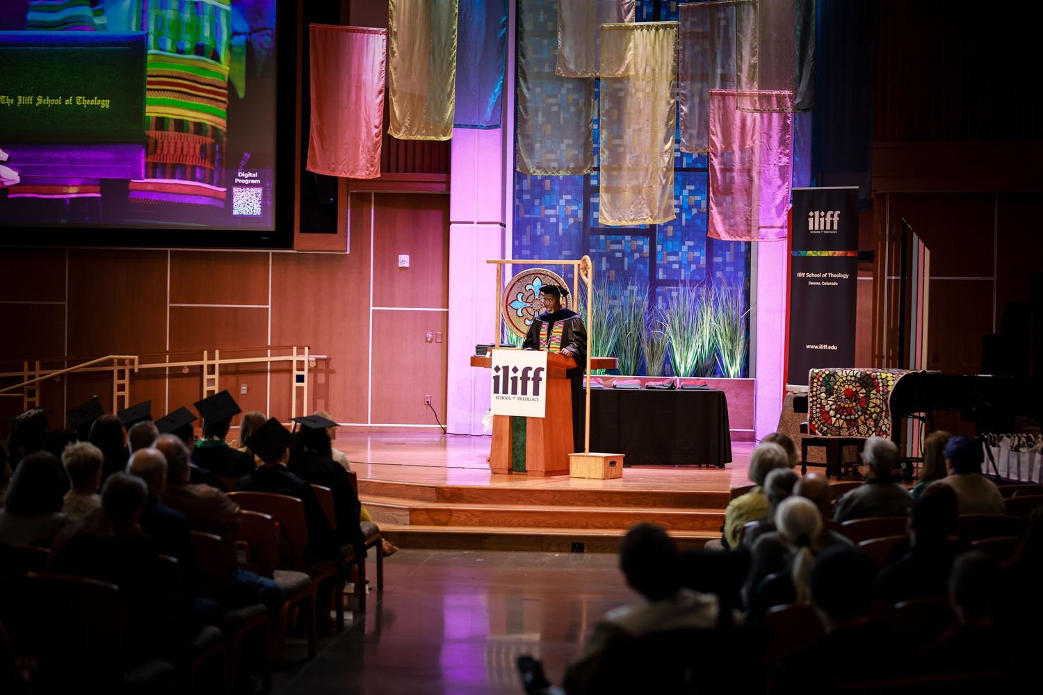 Speaker addresses a large audience at an ILIFF School of Theology event with colorful fabric backdrops.