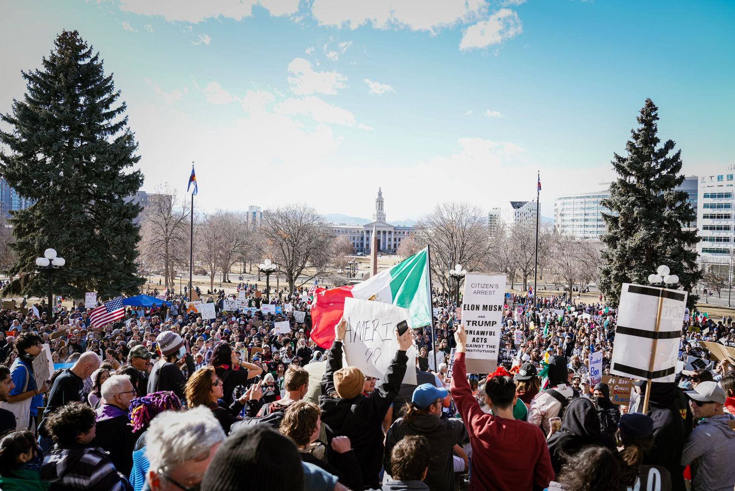 A large diverse crowd rallies outdoors holding signs and a Mexican flag at a public protest.