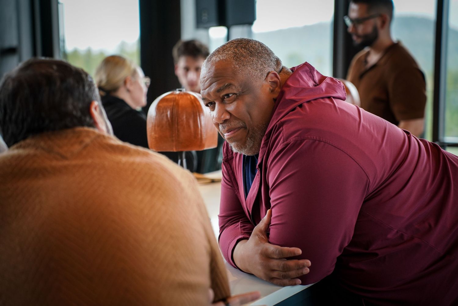 Man in maroon hoodie leaning forward engaged in conversation at table.