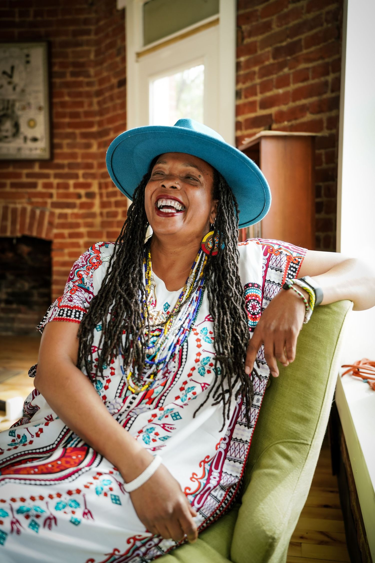 Joyful woman in blue hat, colorful African print dress, and beaded jewelry laughing while seated on green chair.