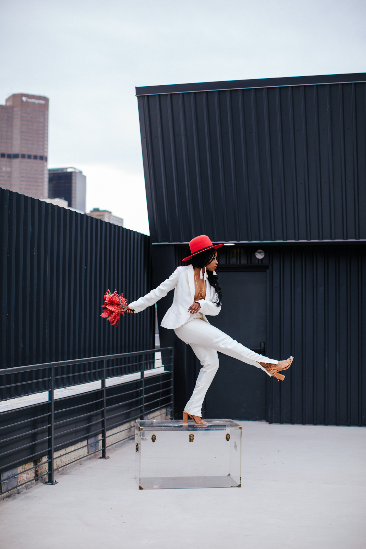 Stylish Black woman in white suit and red hat posing on rooftop with red flowers.