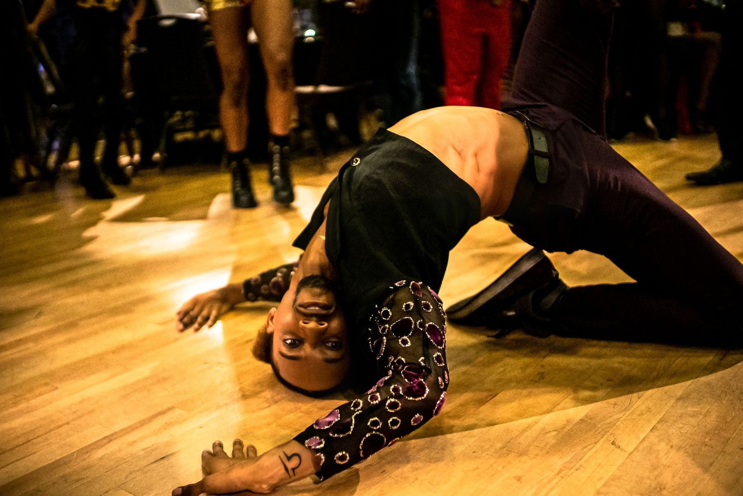 Dancer in patterned outfit performing a dramatic floor move on a wooden dance floor.