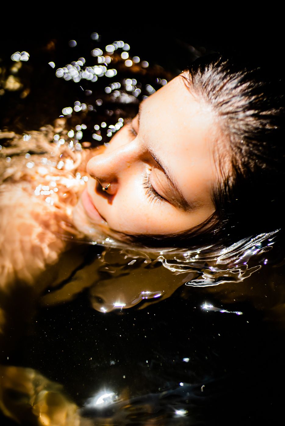 A woman with closed eyes floating peacefully in dark water with bokeh light reflections.