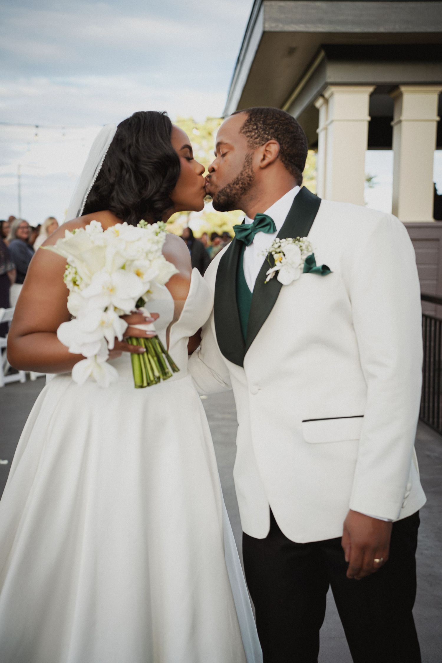 A bride and groom share a kiss at their wedding, she holds white florals, he wears a white tuxedo with green bow tie.
