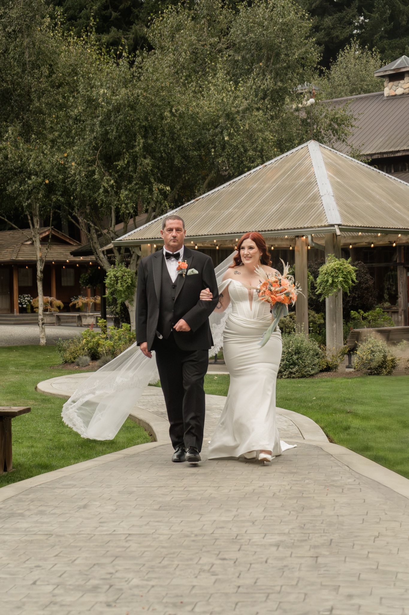 Bride in fitted white gown with billowing veil walking alongside suited escort at outdoor wedding venue.