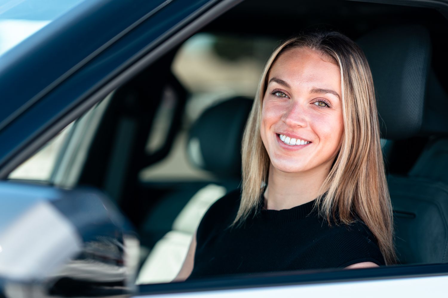 Smiling young blonde woman sitting in driver seat of a car, looking out the open window on a sunny day.