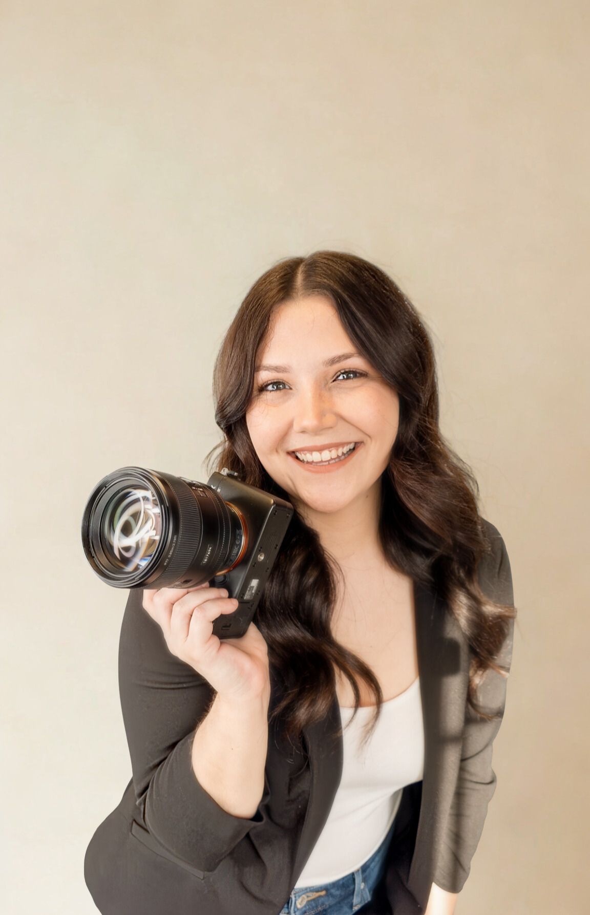 Smiling female photographer holds professional camera in dark blazer against neutral background.
