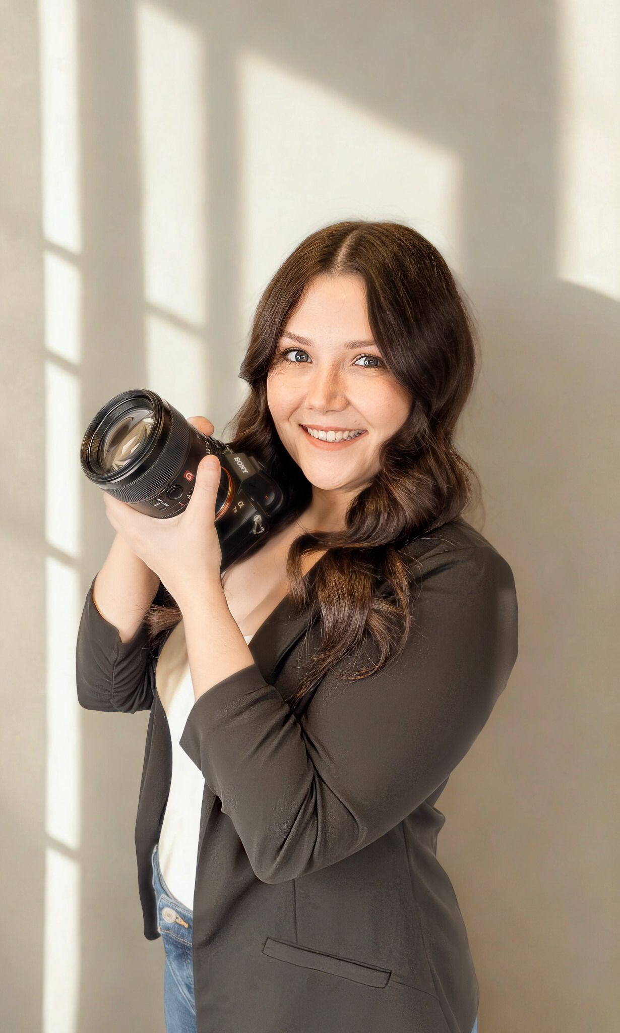 Professional female photographer smiles holding DSLR camera in olive blazer by sunlit window.