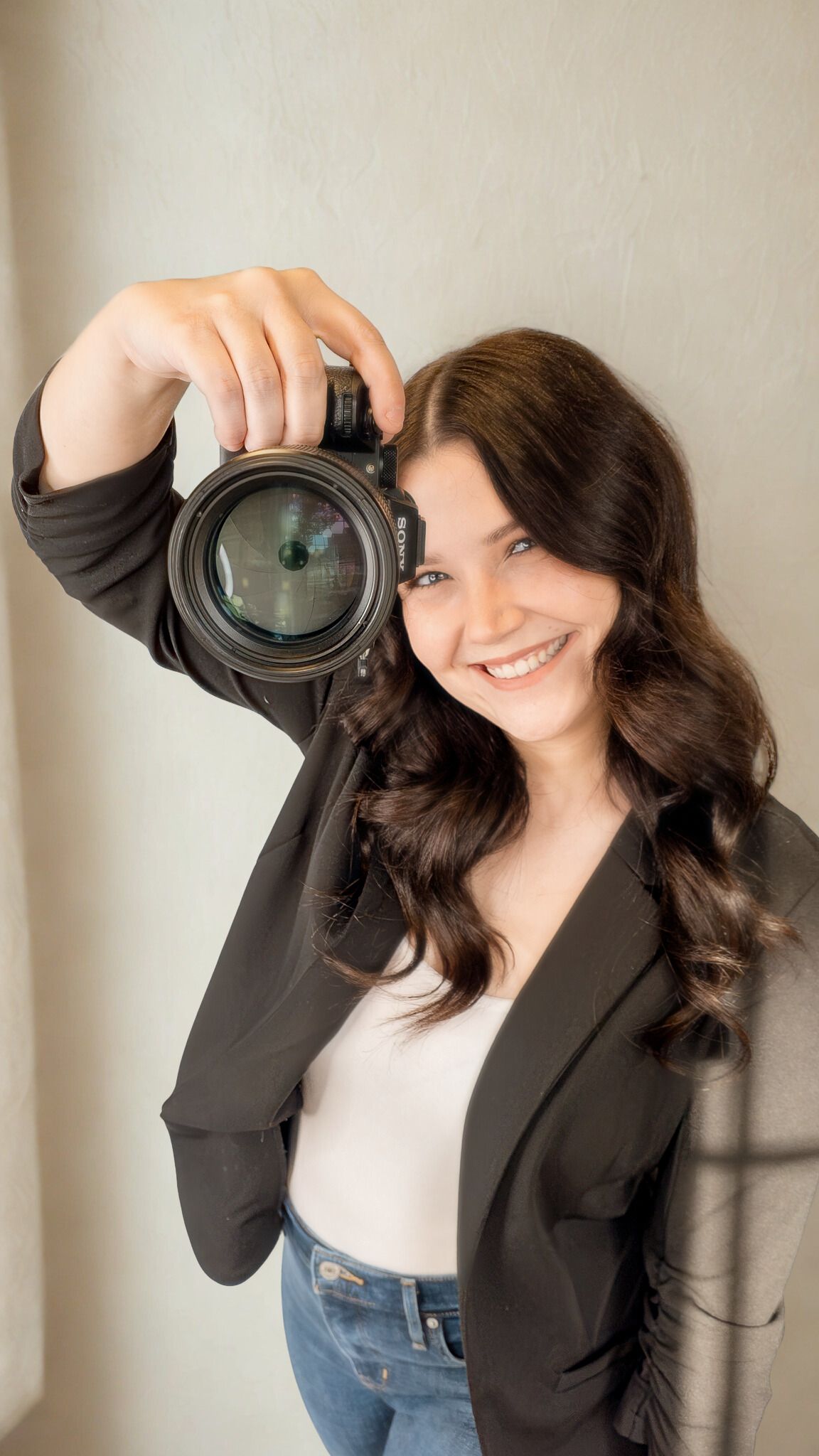 Smiling young woman holding a DSLR camera up, wearing a black blazer and jeans against a light wall.