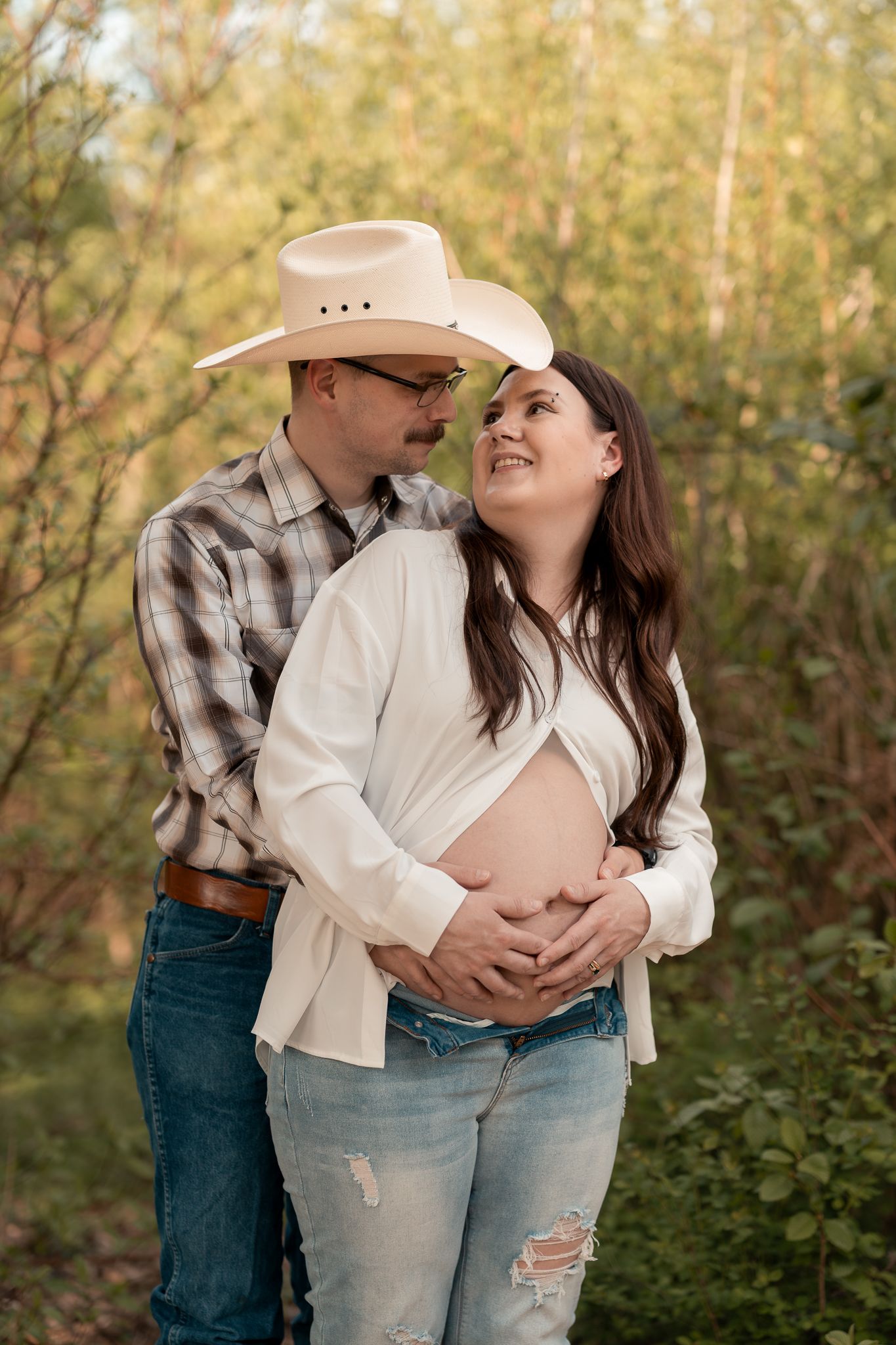 Couple sharing a tender moment outdoors, man in cowboy hat embracing pregnant woman in nature.
