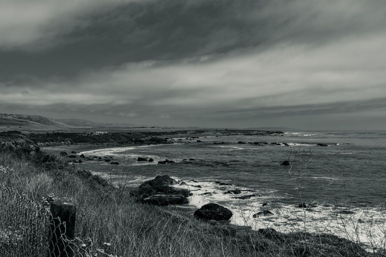 Black and white coastal scene featuring rocky shoreline, crashing waves, and stormy overcast skies along rugged coast.