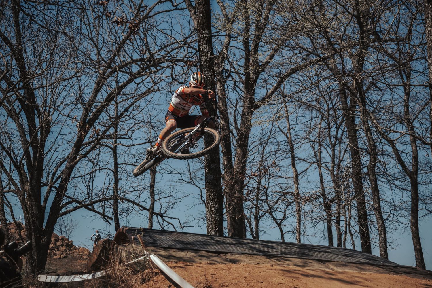 Mountain biker performing a big jump off a wooden ramp feature with bare trees in background.