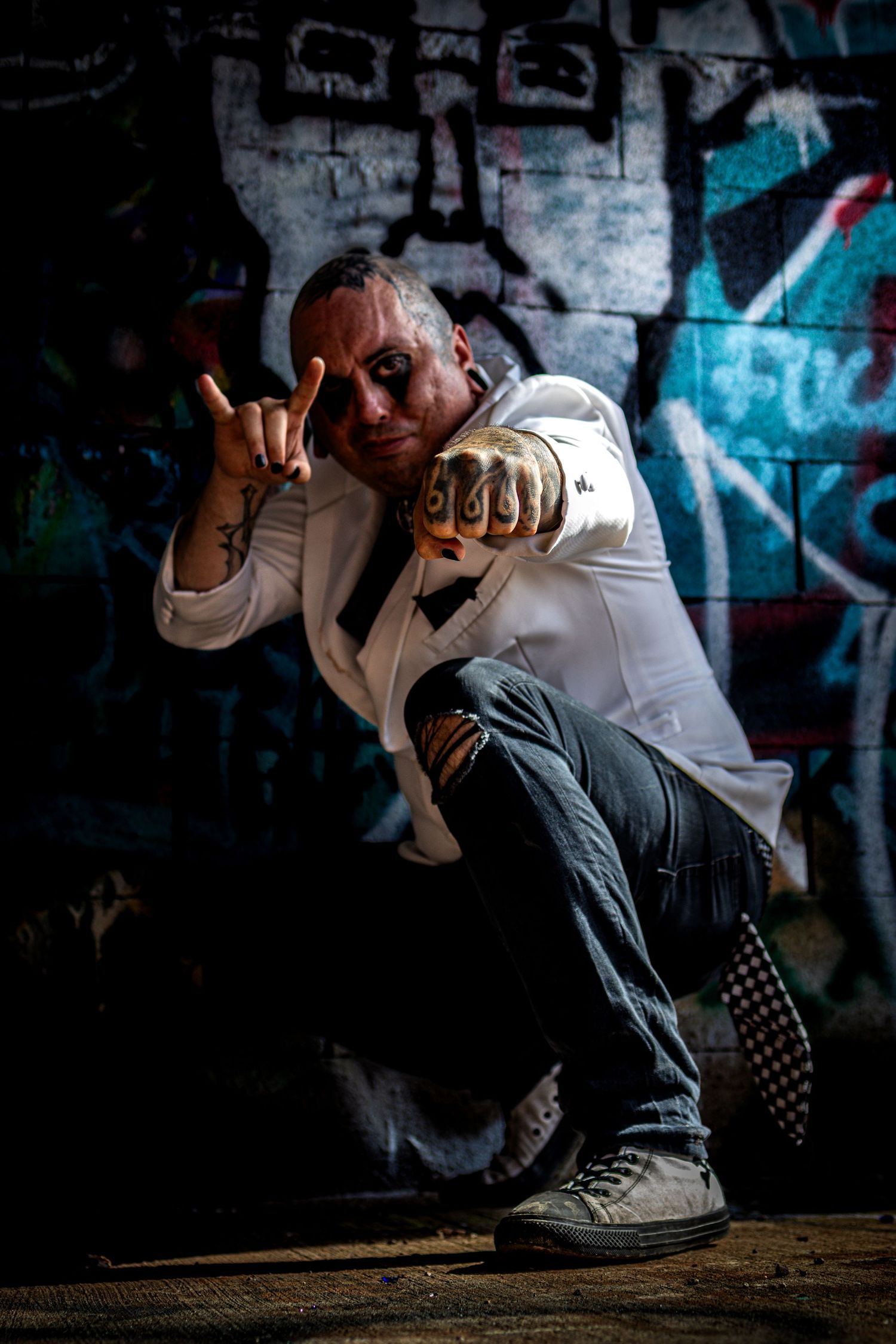 Tattooed man in white shirt crouching against graffiti wall making hand gesture in dramatic lighting.