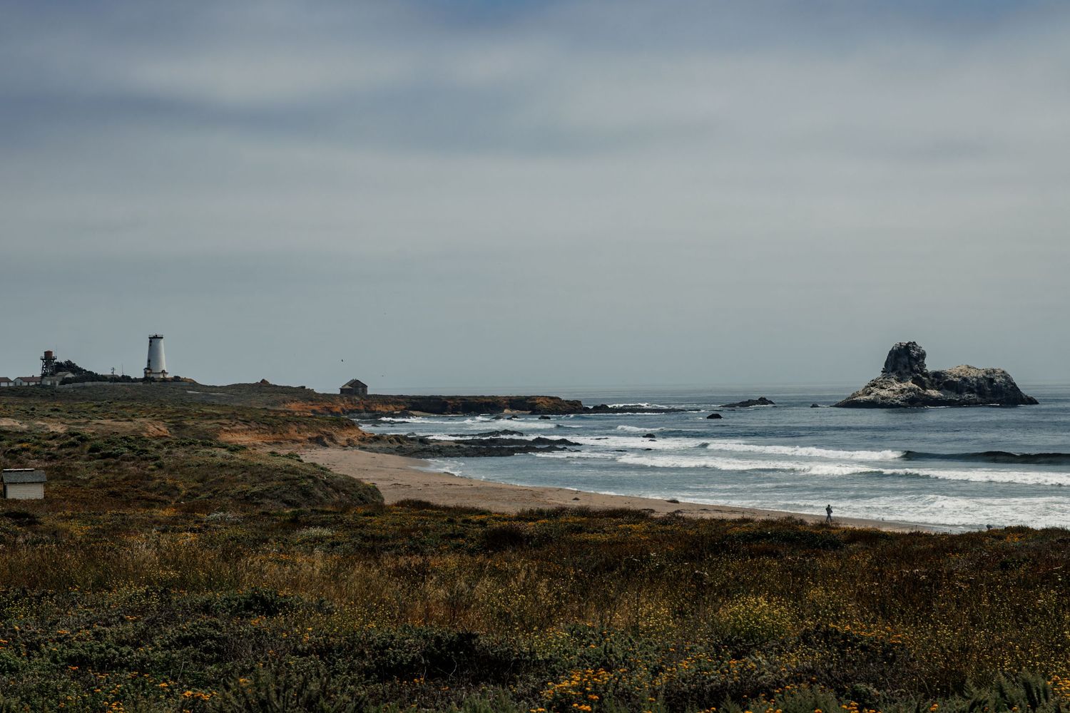 A rocky coastline with a distant lighthouse and sea stacks under an overcast sky near the ocean.