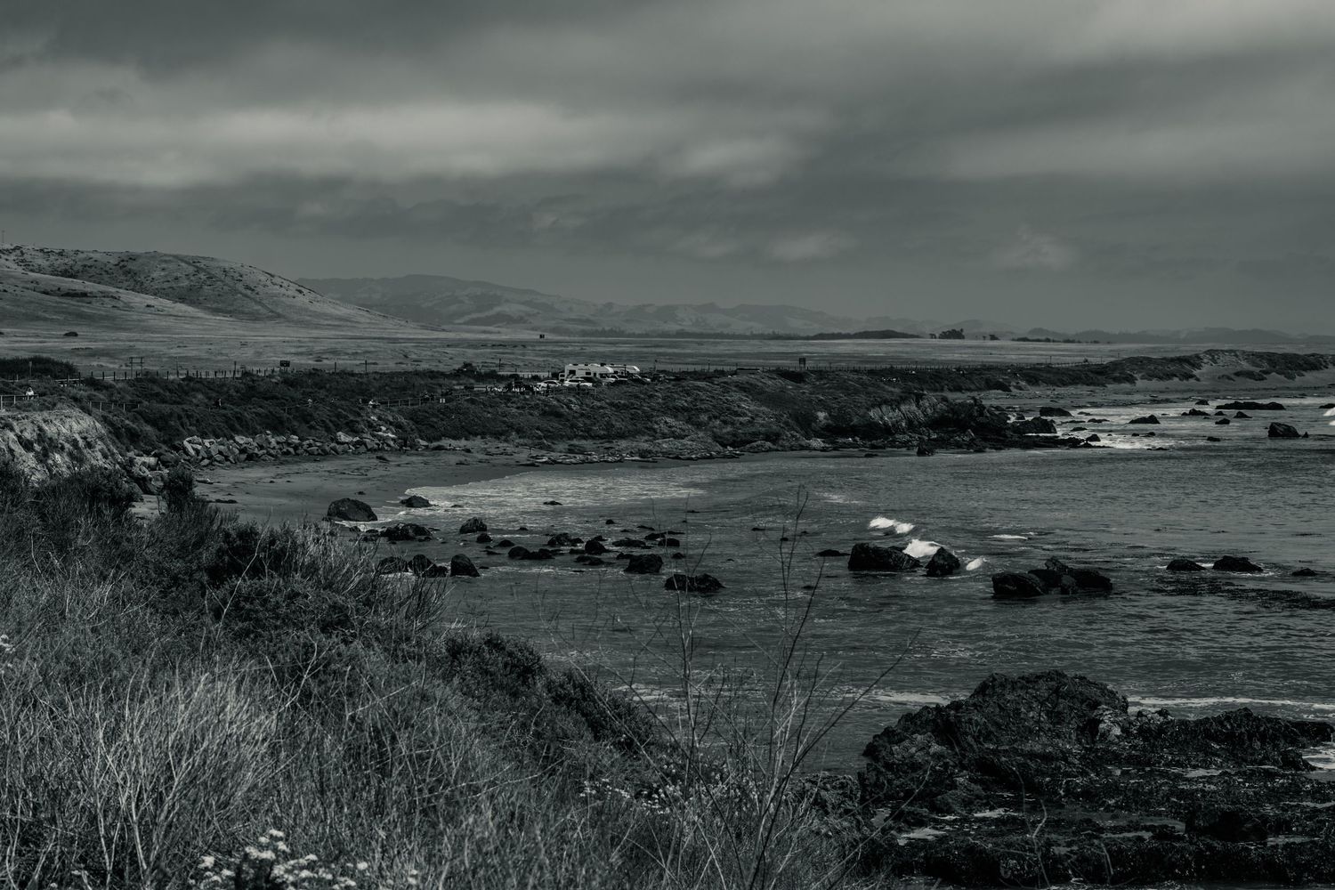 Black and white panoramic coastal landscape with scattered rocks, grassy cliffs, and distant village under cloudy sky.