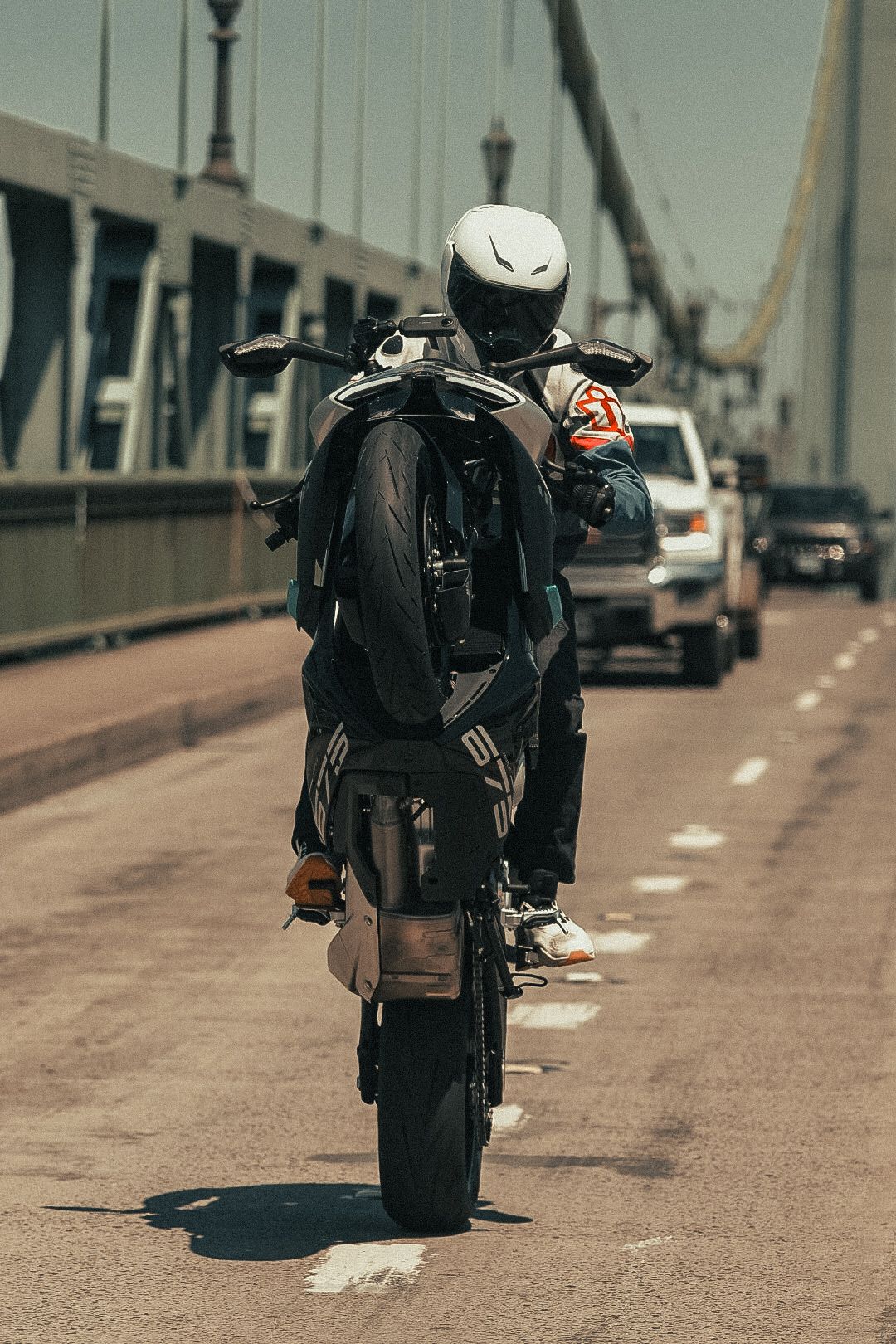 Motorcyclist performing a wheelie on a bridge with traffic in the background on a sunny day.