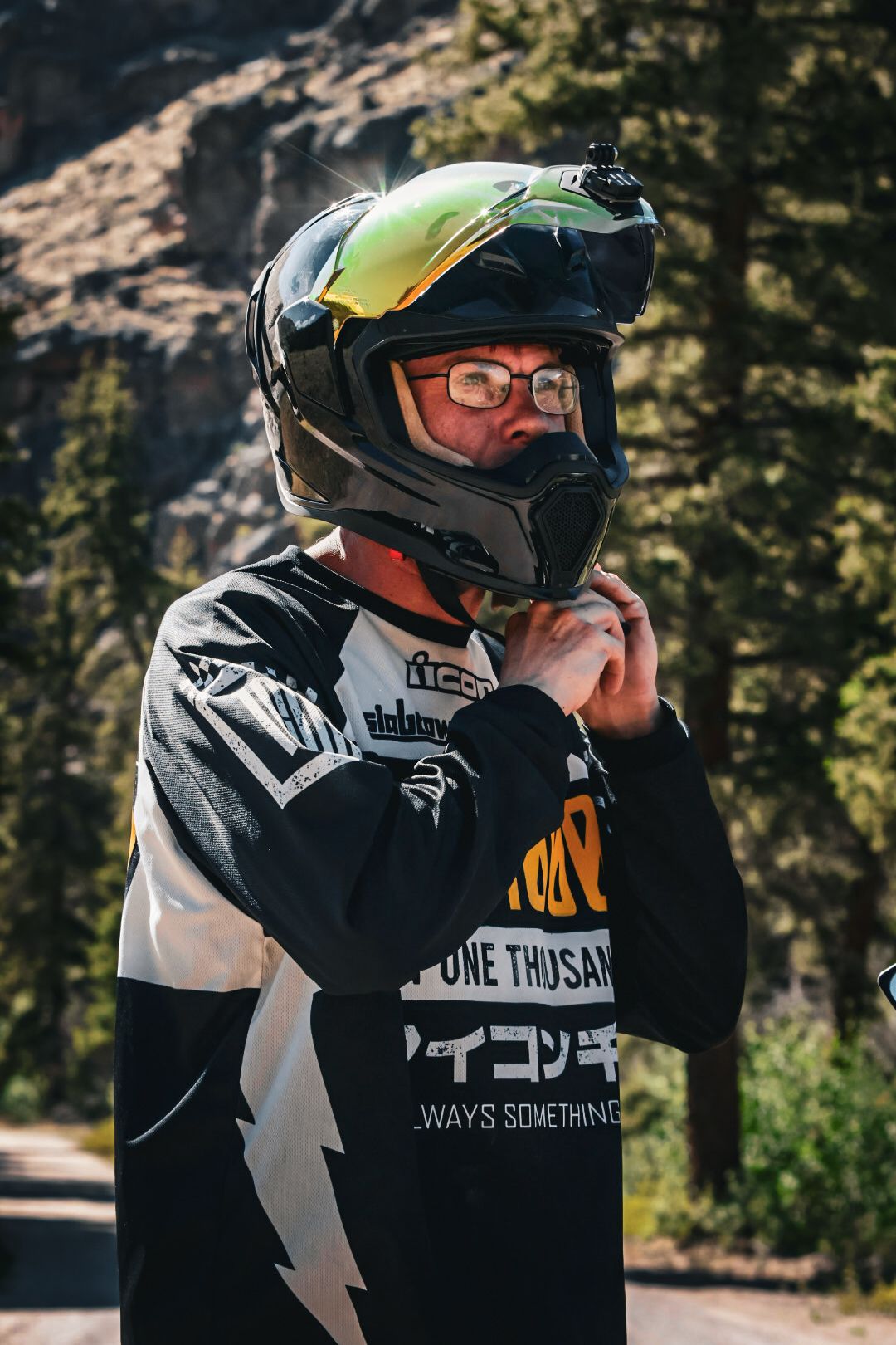 Motorcyclist adjusting black helmet with yellow visor outdoors in mountain forest setting.