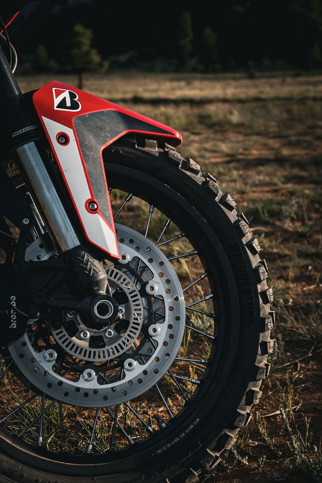 Close-up of a dirt bike front wheel with Bridgestone knobby tire and red fender in field.