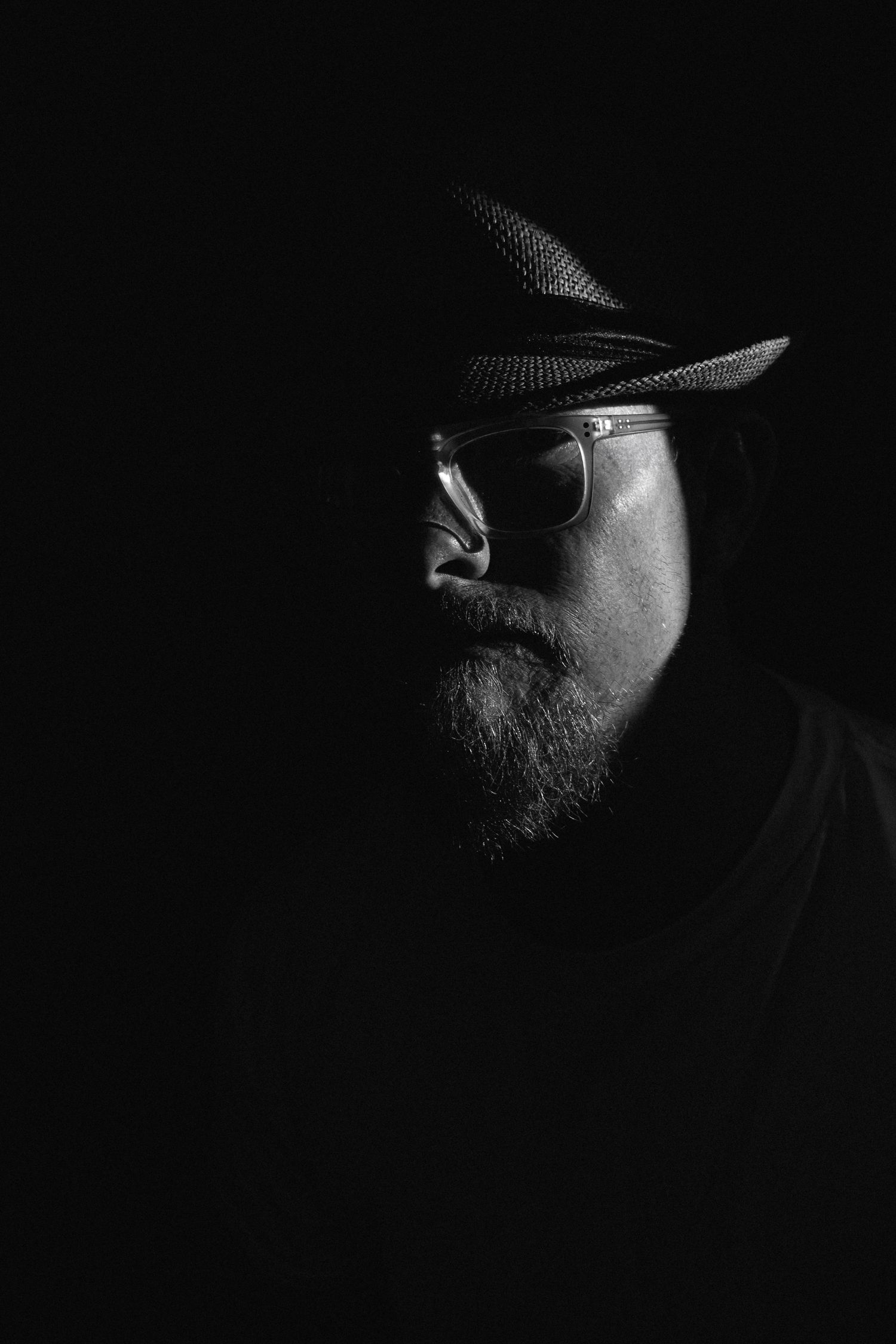 Dramatic black and white portrait of bearded man wearing fedora hat and glasses against dark background.