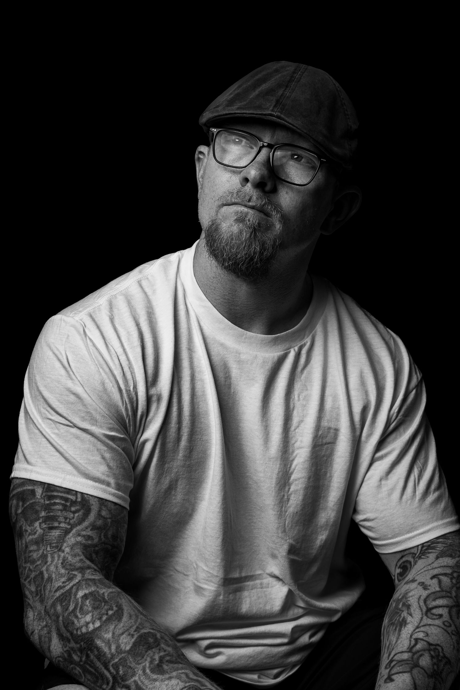 Black and white portrait of tattooed bearded man wearing cap and glasses against dark background.