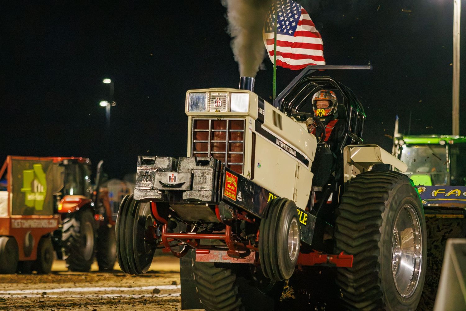 A white tractor with an American flag competes in a nighttime tractor pull event, emitting black smoke.