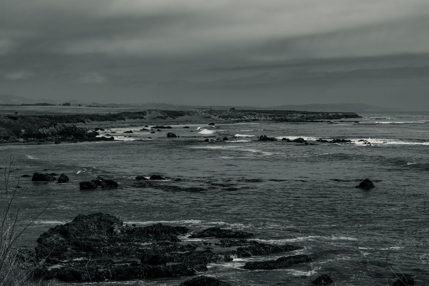 Dark moody black and white seascape with rocky coastline and dramatic storm clouds over rough ocean waters.