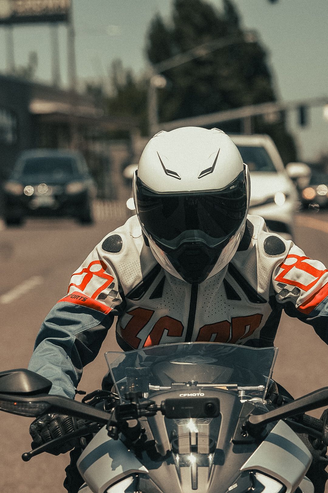 Motorcyclist in white helmet and racing jacket stopped at a busy urban intersection with traffic.