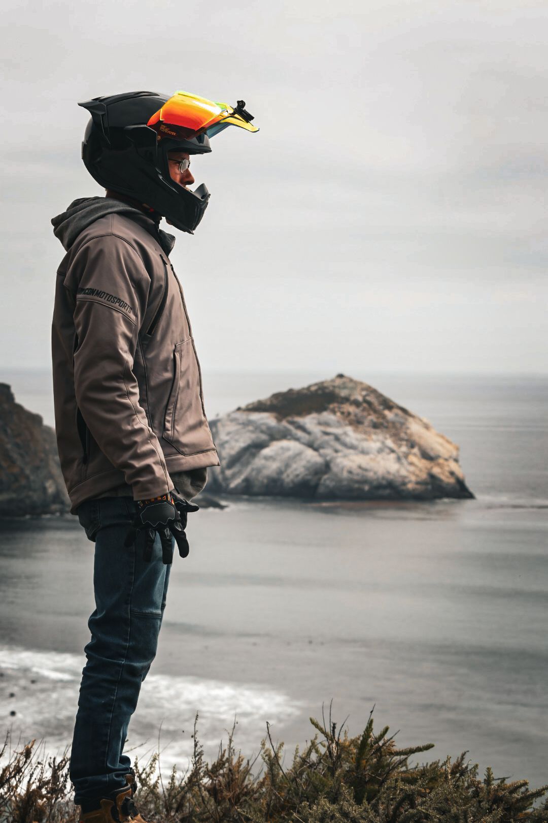 A motorcyclist in a black helmet with orange goggles stands on a coastal cliff overlooking the sea.