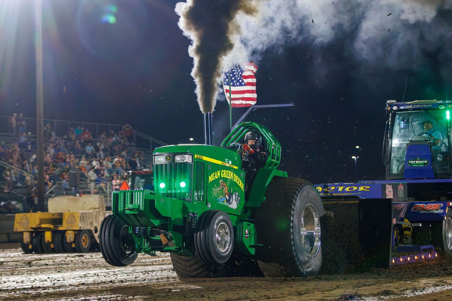 A green John Deere tractor with an American flag powers through a dirt track during a nighttime tractor pull competition.