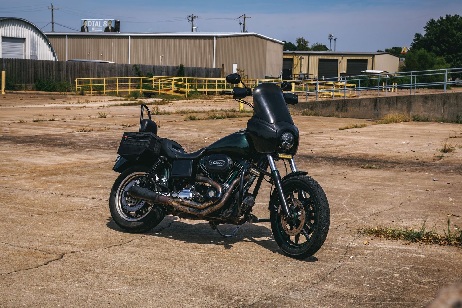 Black Harley-Davidson motorcycle with saddlebags parked in an industrial lot with yellow fence and buildings.