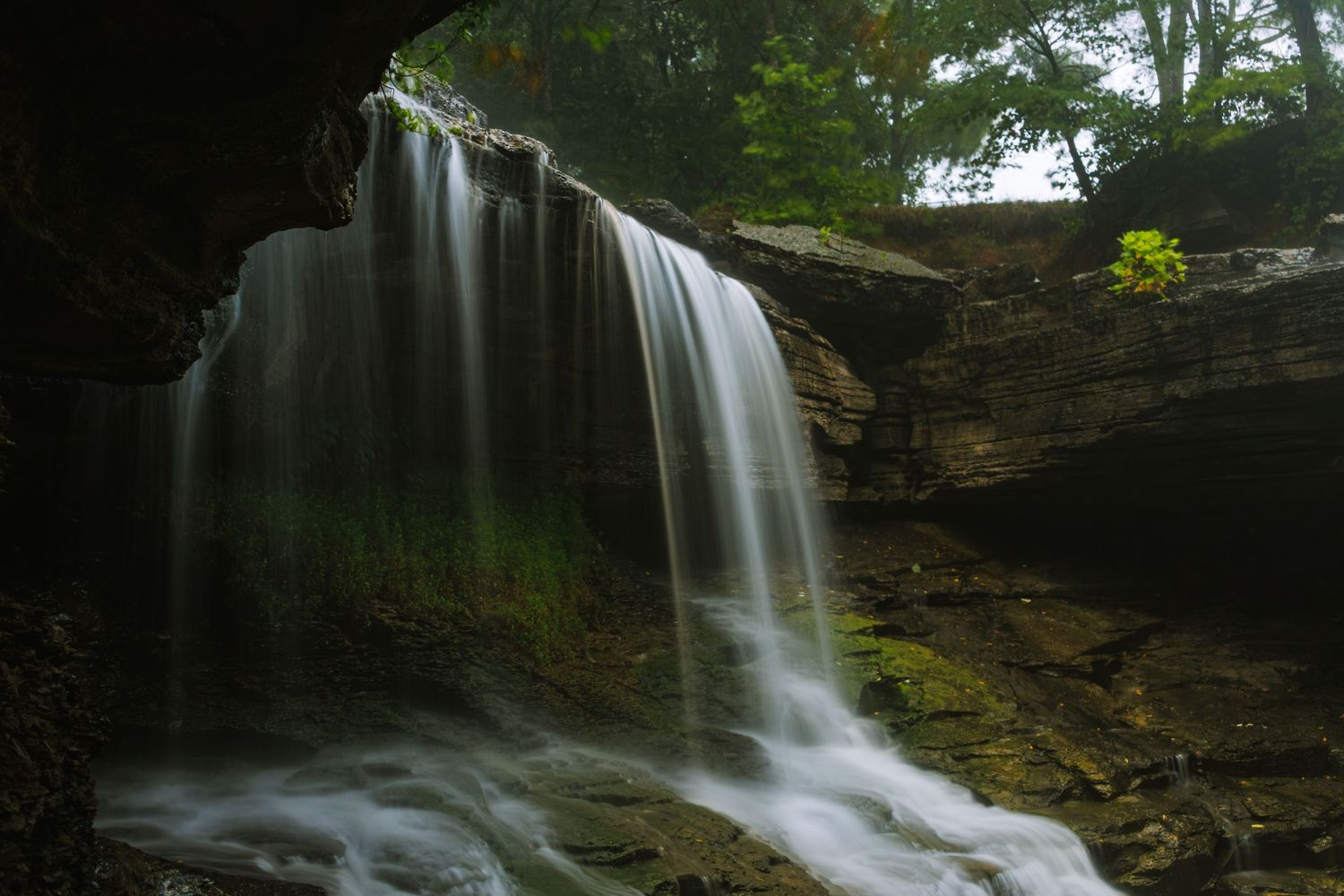 A stunning waterfall cascades over mossy rocks viewed from inside a cave in a lush forest setting.