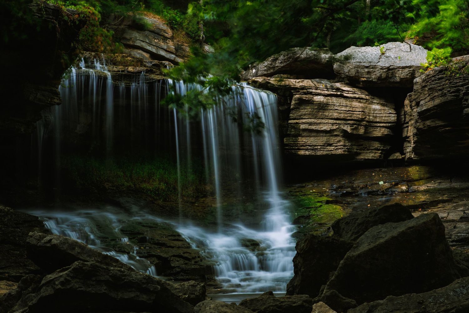 A serene waterfall flows over layered rock formations surrounded by lush green forest and boulders.