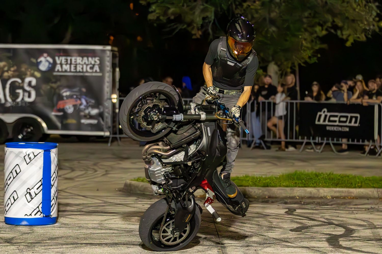 Motorcycle stunt performer doing a dramatic wheelie near a barrel at a nighttime event.