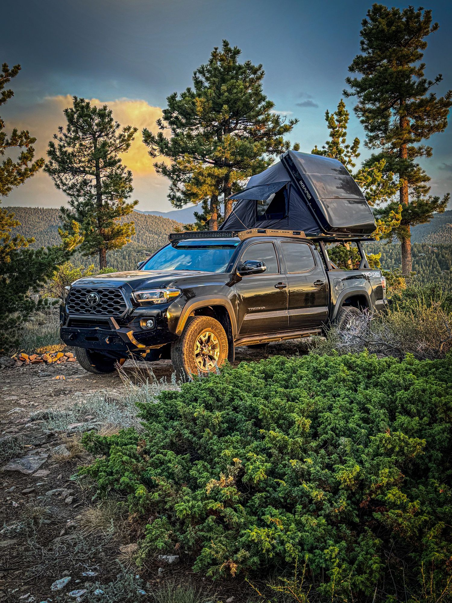 Black Toyota Tacoma with rooftop tent parked on rocky mountain terrain at dusk.