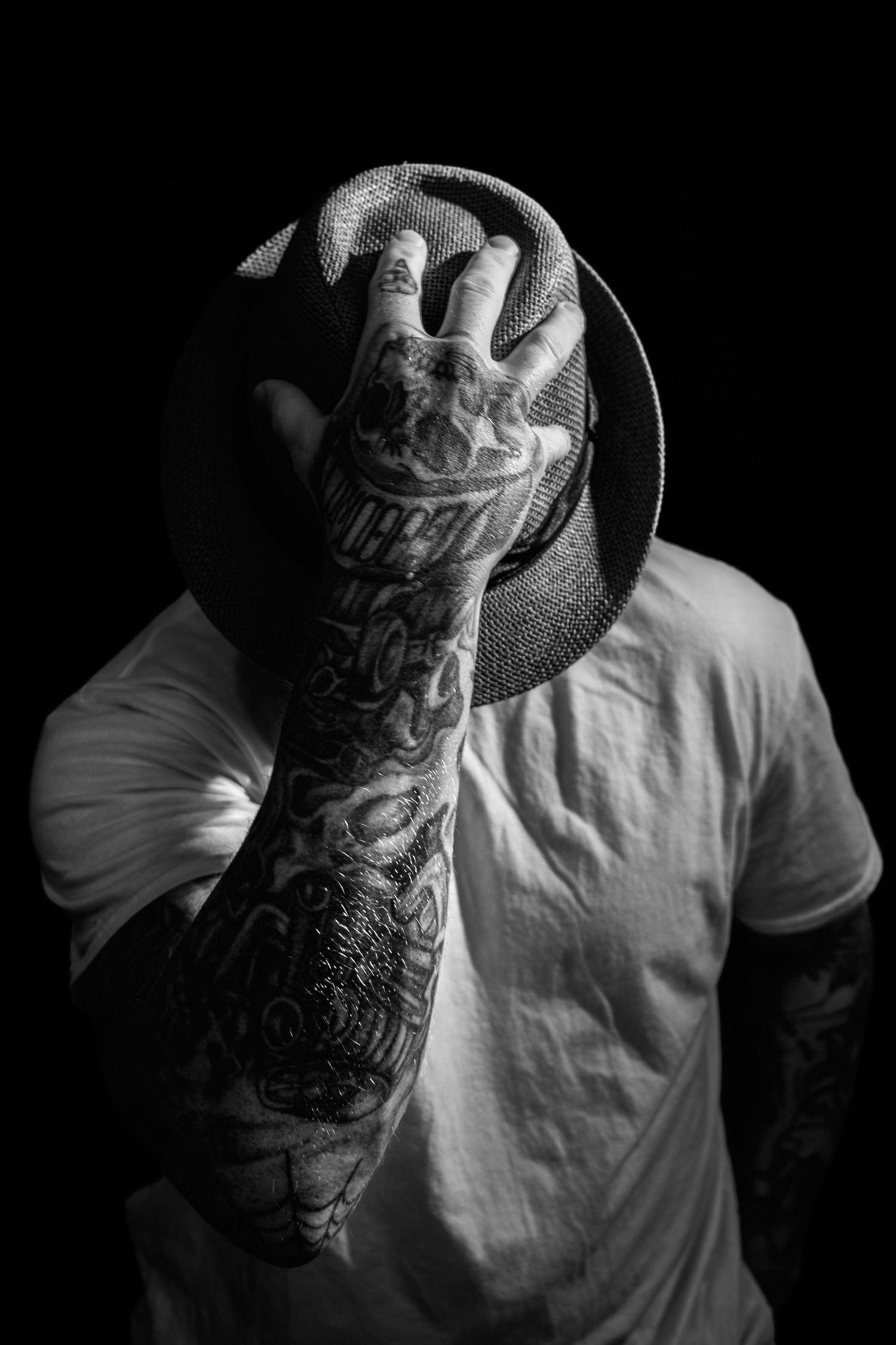 Black and white portrait of a tattooed man hiding his face behind a hat against dark background.